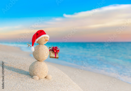 Christmas Snowman in Santa hat with gift at sunset beach