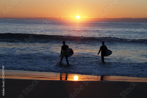 surfers walking into ocean as sun breaks