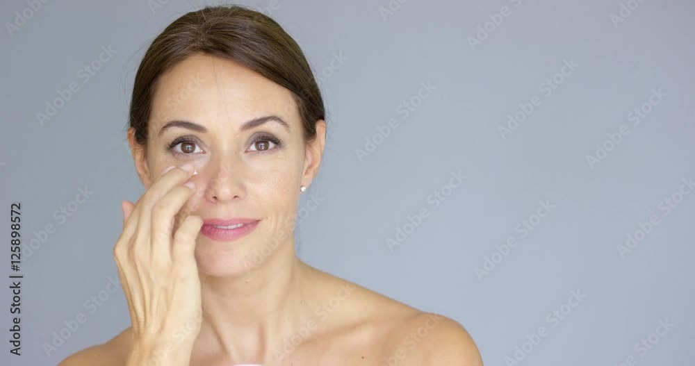 Beautiful young woman applying face cream to her cheek bone in a beauty skincare or cosmetics concept on grey with copy space Beautiful young woman applying face cream to her cheek bone in a beauty skincare or cosmetics concept on grey with copy space