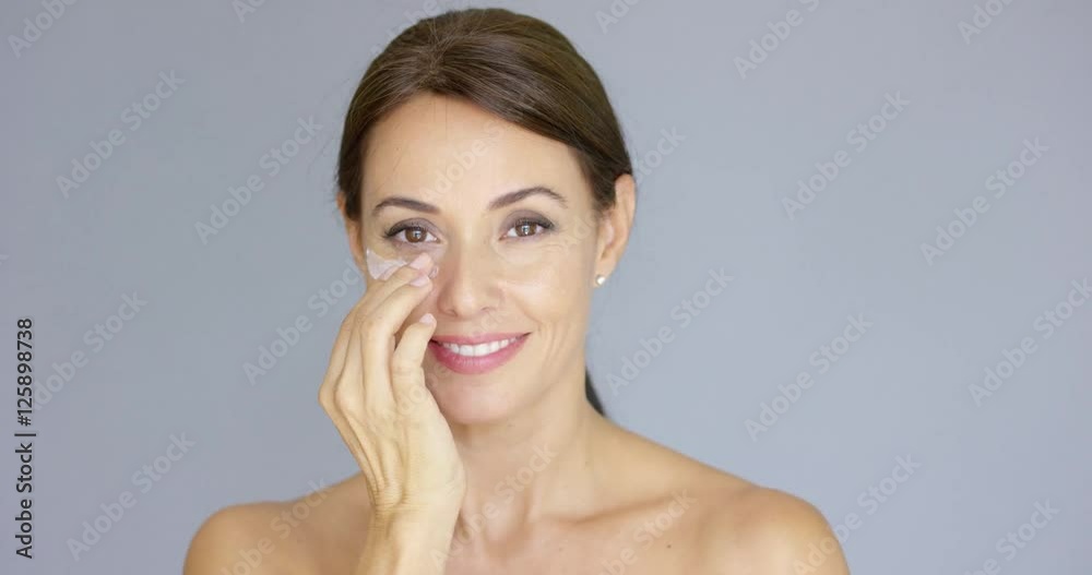 Gorgeous young woman applying face cream to her cheek with her fingertips with a smile in a skincare and beauty concept  close up head and shoulders over grey.
