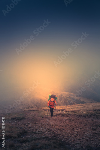 Lonely hiker with backpack walking along the trail on the mountain top at foggy day time. Travel lifestyle concept.