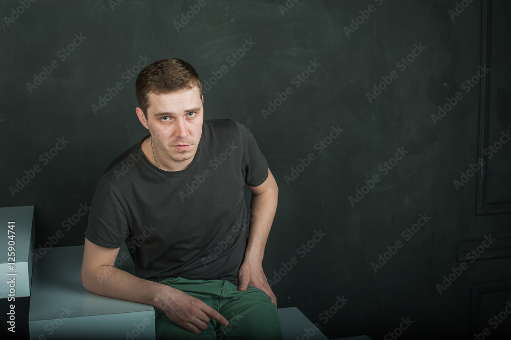 studio photography young brutal guy. man in T-shirt on a background of black wall on the white steps. sitting on the steps, looking at the camera studio photography young brutal guy. man in T-shirt on a background of black wall on the white steps. sitting on the steps, looking at the camera