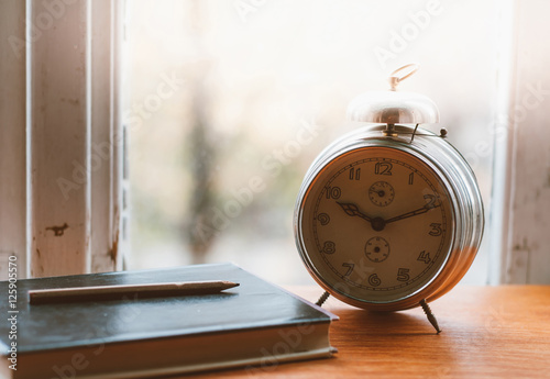 Vintage book and metal analog alarm clock with Arabic numbers and wind-up mechanism, sitting on a wooden bench with retro background. Time is now, time is money, old times concept. Copypace included.