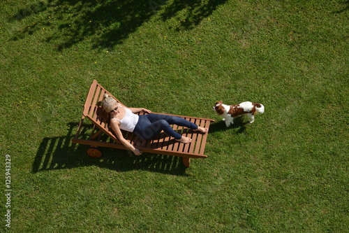Canvas Print Woman resting on a wooden deckchair on a garden lawn while her dog (Cavalier Kin