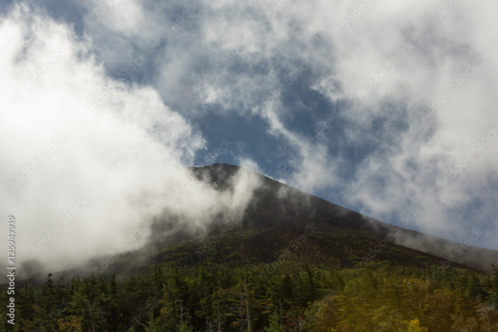 Mount Fuji, Japan - September 27, 2016: Dark Mount Fuji summit under white clouds as seen from Walk-up Station Five. Forests in foreground.