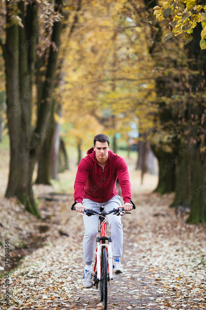 Autumn outdoors. Young serious handsome guy with earphones rides his bike in a city park and listening to music.