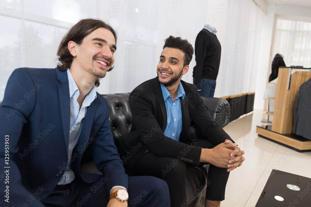 Two Young Handsome Business Man Sitting On Coach In Retail Store Speaking Shopping, Elegant Businessman Choosing Formal Wear Two Young Handsome Business Man Sitting On Coach In Retail Store Speaking Shopping, Elegant Businessman Choosing Formal Wear