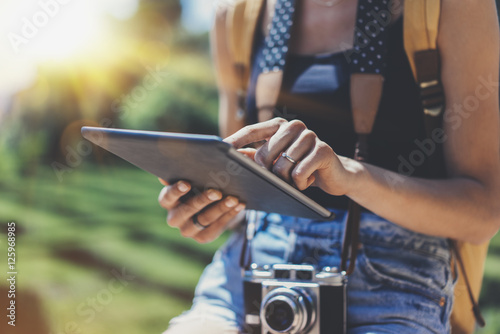 Hipster girl with backpack and vintage photo camera using tablet computer close up, copy space of blank empty mockup, view tourist hands holding gadget and texting message on background sun flare