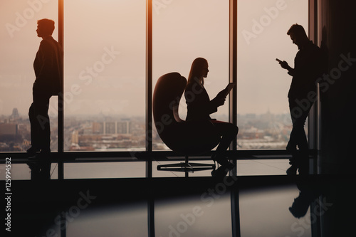 Silhouettes of business people two man and one woman resting and waiting for the meeting near big window in luxury office interior with reflections, sunset, high floor, winter cityscape outside