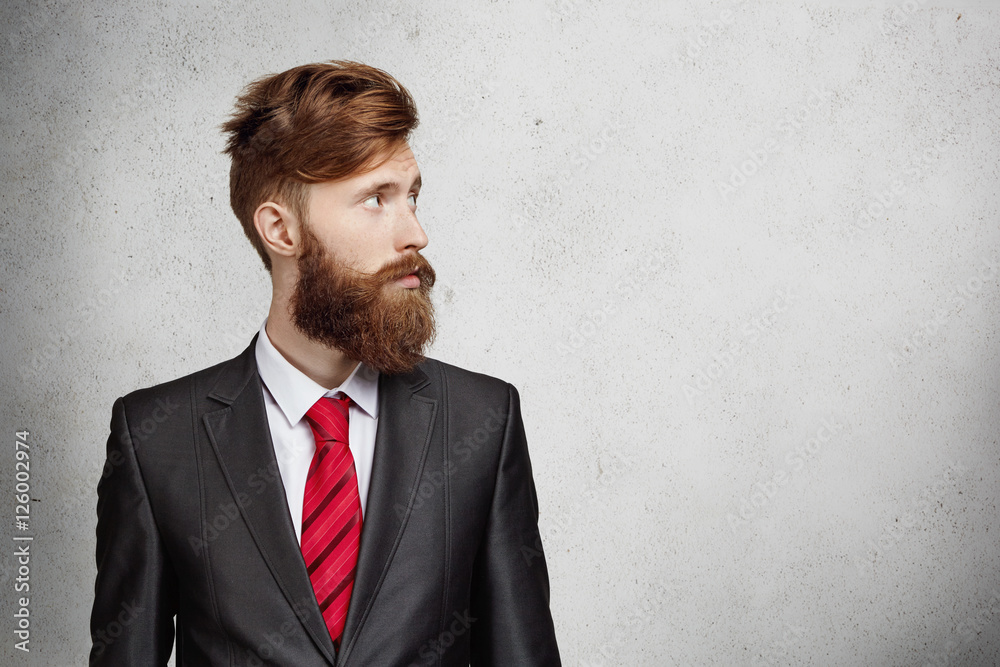 Young successful businessman dressed in elegant suit with red tie looking at blank wall with copy space for your content. Stylish bearded entrepreneur making up his mind before serious negotiations Young successful businessman dressed in elegant suit with red tie looking at blank wall with copy space for your content. Stylish bearded entrepreneur making up his mind before serious negotiations