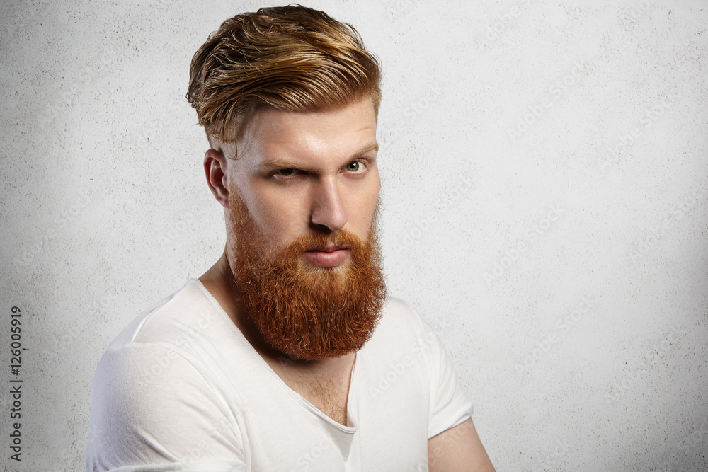 Studio shot of handsome fashionable man with long red beard and trendy hairstyle having serious and displeased facial expression scowling and frowning while standing against concrete wall background Studio shot of handsome fashionable man with long red beard and trendy hairstyle having serious and displeased facial expression scowling and frowning while standing against concrete wall background