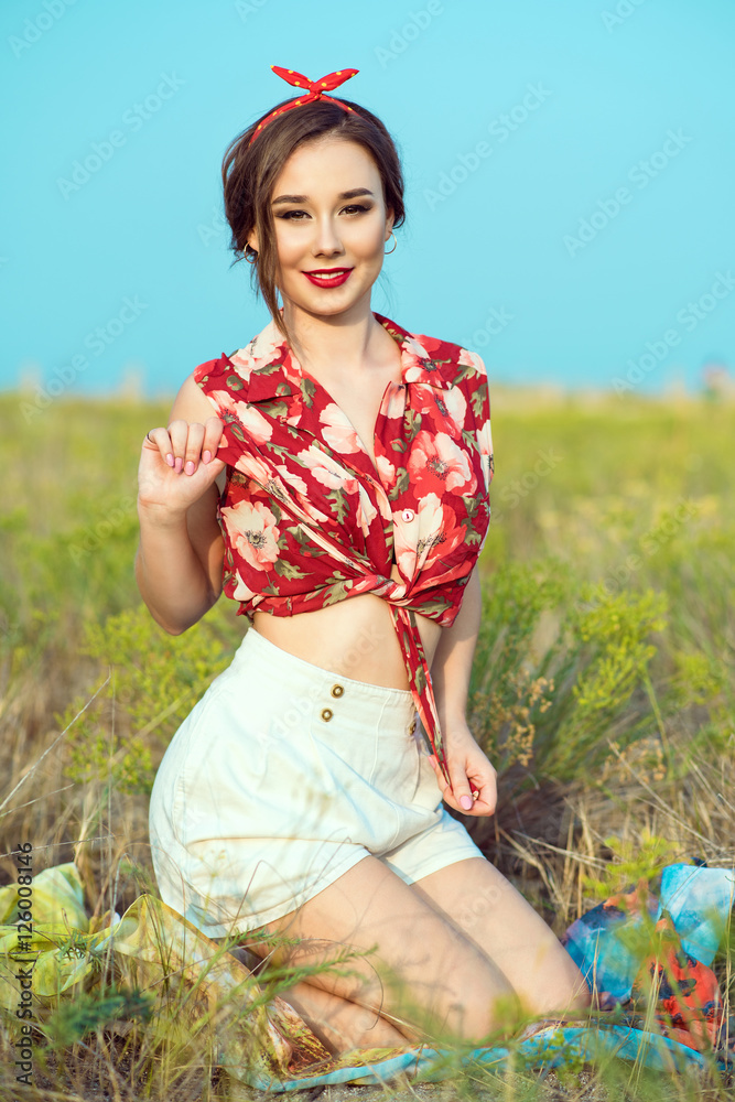 Beautiful young girl with perfect tempting smile wearing red tied up blouse,  white shorts and headband sitting in the field. Pin-up style. Outdoor shot.  Stock-Foto | Adobe Stock