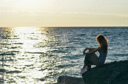young red-haired woman  sitting on the rocks and watching at the sunset on the sea