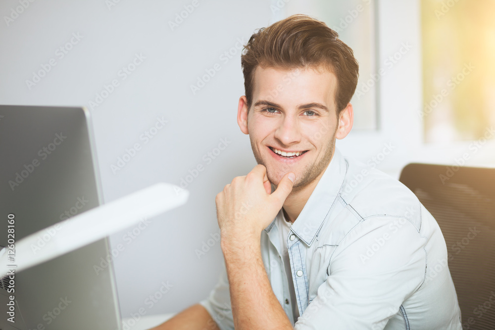 A young programmer is looking at the camera. The graphic designer is thinking about the future projects. The young guy is sitting at the computer in the office with the windows behind his back A young programmer is looking at the camera. The graphic designer is thinking about the future projects. The young guy is sitting at the computer in the office with the windows behind his back
