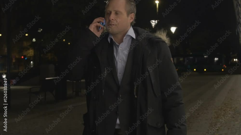 Businessman talking on smartphone and walking towards camera in the evening. Wearing suit jacket and winter jacket outdoors