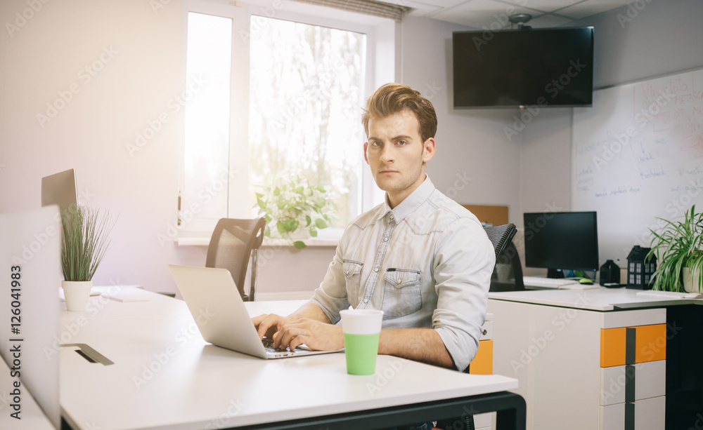 The young guy is sitting at the computer in the office with the windows behind his back