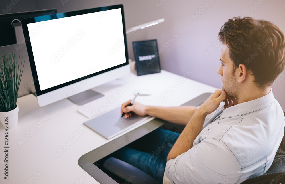 Young worker sitting in an office at the computer. Freelancer in a blue shirt. The designer sits in front of window in the workplace. Young worker sitting in an office at the computer. Freelancer in a blue shirt. The designer sits in front of window in the workplace.