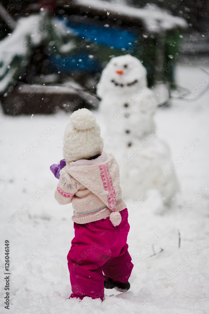 A toddler girl in a pink snowsuit with wooly hat toque builds a large snowman with carrot nose and smiling face in backyard with snowflakes snow falling in winter at Christmas A toddler girl in a pink snowsuit with wooly hat toque builds a large snowman with carrot nose and smiling face in backyard with snowflakes snow falling in winter at Christmas