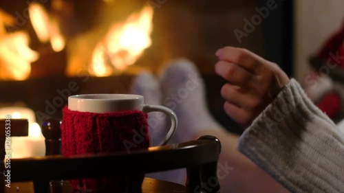 Woman is sitting with cup of hot drink and book near the fireplace