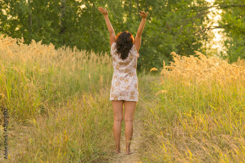 Adult brunette woman in a dress with hands raised up standing on a forest path, view from the back