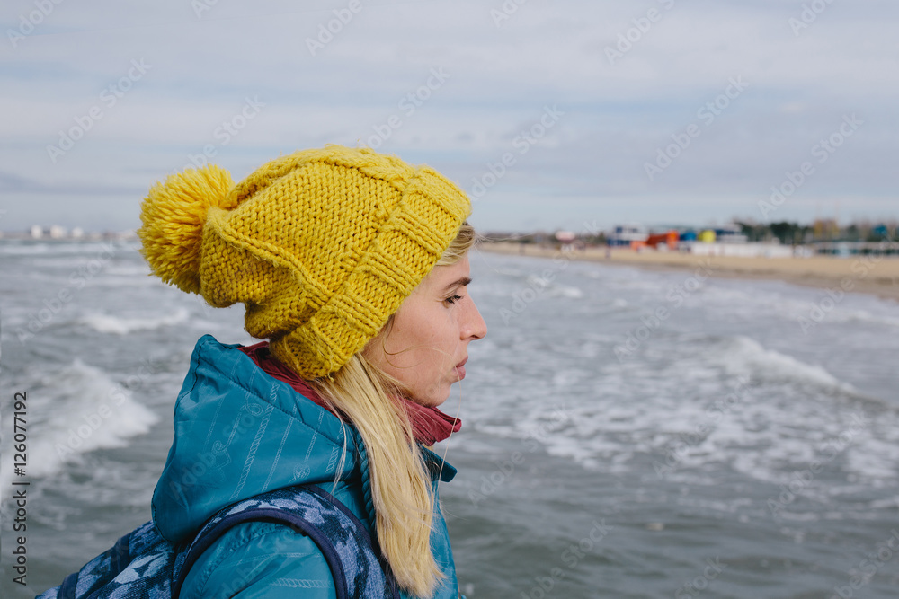 portrait of young woman in a blue jacket and yellow knitted cap on the background stormy sea