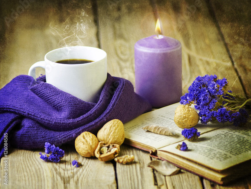 Hot coffee wrapped with cozy wool scarf places with purple candle ,vintage book ,dry flowers and walnut on rustic wooden table.