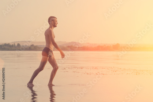 Adult man with a mohawk on his head and black shorts walking on water at sunset or sunrise
