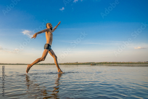 Adult man with a mohawk on his head and black shorts running on water against the backdrop of blue sky with clouds