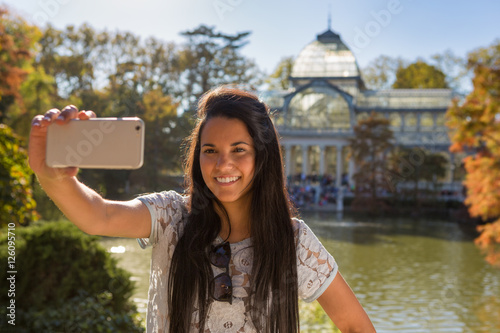 Young female traveler taking a self portrayed (selfie) in the Crystal Palace (Retiro park) of Madrid city in a sunny autumn day.
