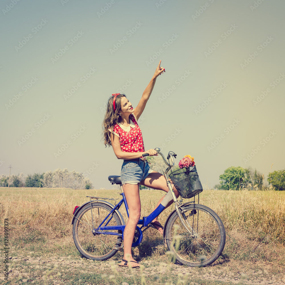 Beautiful pinup girl having fun cycling in fields under bright blue summer sky and pointing up on summer outdoors copy space background
