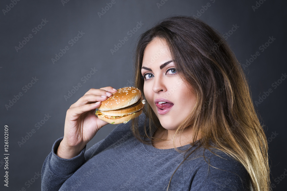 Happy beautiful young plus size model posing with hamburger on a gray studio background, xxl woman eating burger Happy beautiful young plus size model posing with hamburger on a gray studio background, xxl woman eating burger