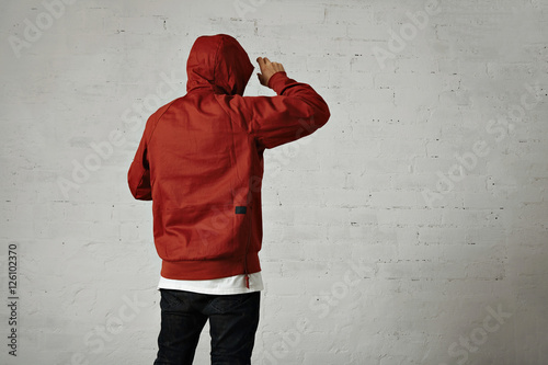 Back shot of a man in black jeans, white t-shirt and red parka putting on his hood against white wall background