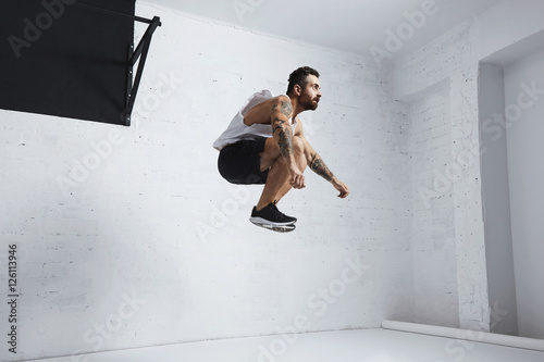 Bearded and tattooed young male athlete shows calisthenic moves, jumping high in air, isolated in white room of fitness center