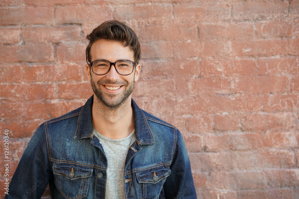 Smiling trendy guy with blue jeans jacket and eyeglasses standing by brick wall Smiling trendy guy with blue jeans jacket and eyeglasses standing by brick wall
