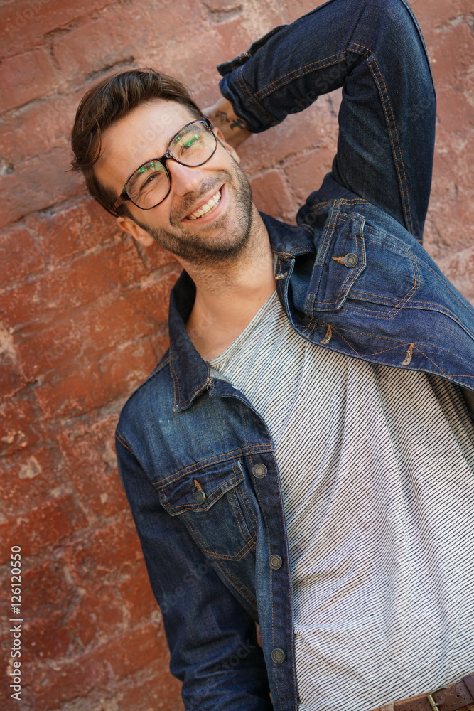 Trendy guy with blue jeans jacket and eyeglasses standing by brick wall Trendy guy with blue jeans jacket and eyeglasses standing by brick wall