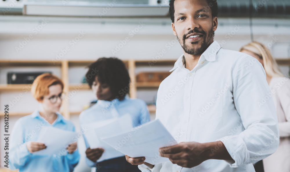 Fototapeta premium Dark skinned entrepreneur holding papers in hands and smiling at the camera.Teamwork concept in modern office. Horizontal,blurred background.