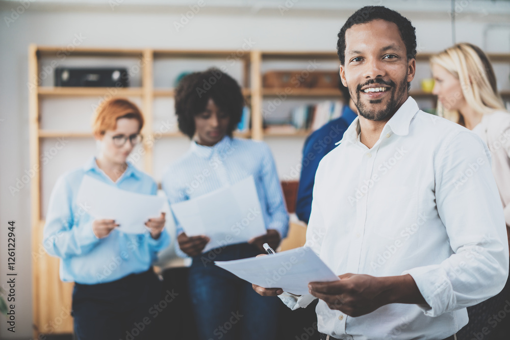 © SFIO CRACHO - African american entrepreneur in white shirt holding papers in hands and smiling at the camera.Teamwork concept in modern office. Horizontal,blurred background.
