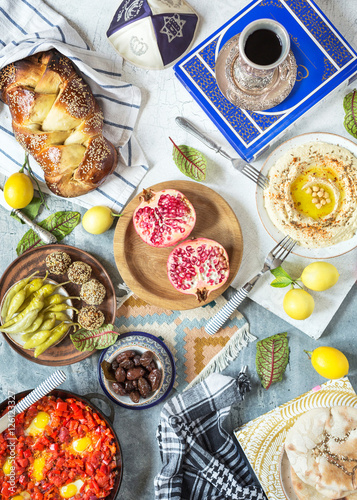 Top view coposition of israel and middle eastern food: ritual goblet of wine, challah and pita bread, hummus, falafel, shakshuka, lemons, pomegranate and olives on rustic table