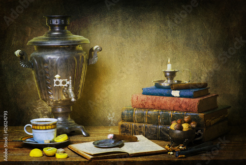 Classic still life with cup of hot tea and some slices of lemon  place together with old books, some nuts,cigar,vintage samovar and silver candle on rustic wooden table.