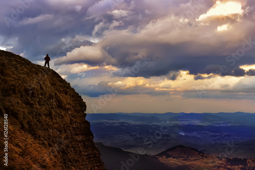 silhouette, figure of a man on a mountain top. In the background is green sloping mountains, low heavy sky with volumetric clouds textured. 