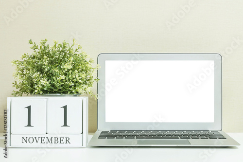 White wooden calendar with black 11 november word with white blank space at center of computer notebook on white wood desk and cream wallpaper textured background , selective focus at the calendar