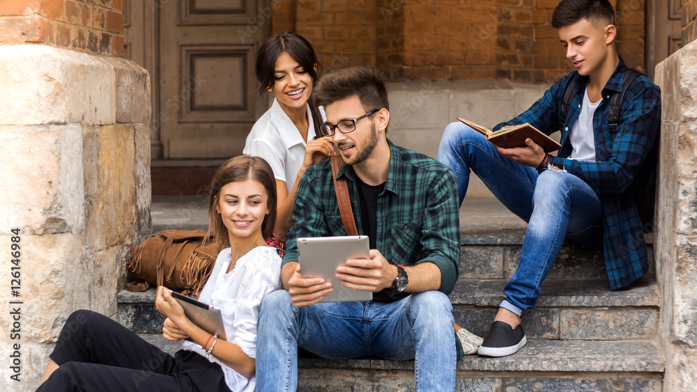 Students life. Low angle view of cheerful young people communicating while holding different gadgets and sitting close to each other on steps.