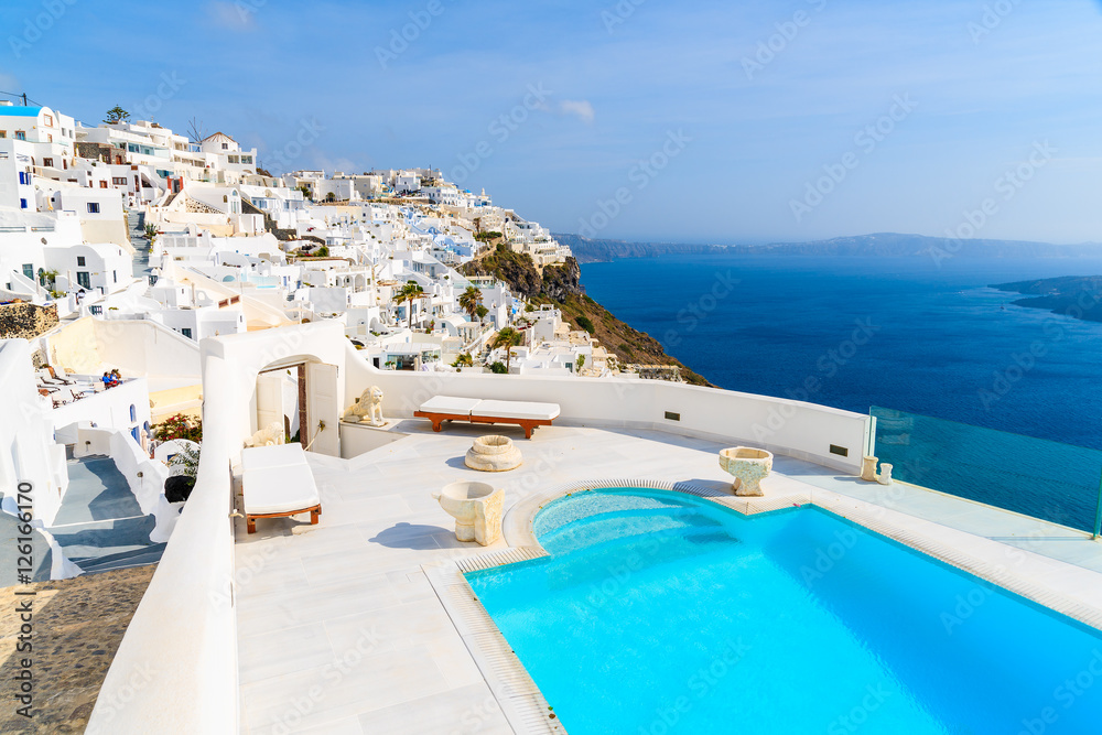 View of caldera and luxury swimming pool in foreground, typical white architecture of Imerovigli village on Santorini island, Greece.