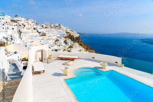 View of caldera and luxury swimming pool in foreground, typical white architecture of Imerovigli village on Santorini island, Greece.