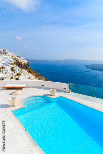 View of caldera and luxury swimming pool in foreground, typical white architecture of Imerovigli village on Santorini island, Greece.