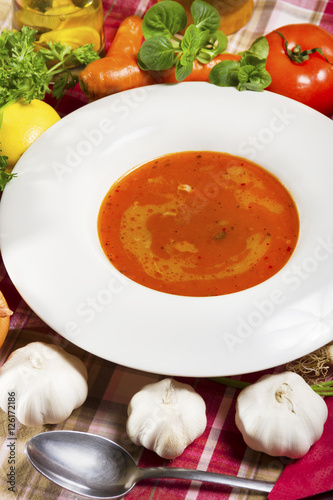Red fish soup in a white plate beside fresh vegetables.