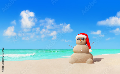 Sandy snowman in Christmas Santa hat and sunglasses at beach