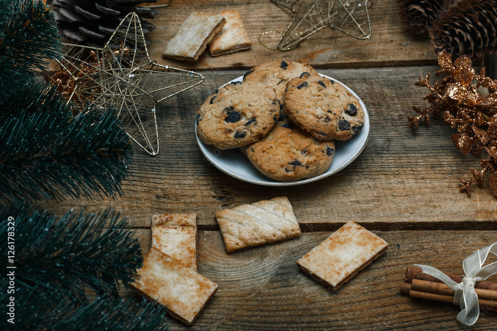 Fototapeta premium Chocolate cookies in a white plate on wooden background. Near cones and cinnamon stick, roughly. View from above. The theme of the new year holiday
