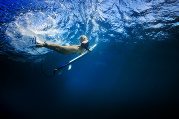 Slim blonde girl in bikini a surfer with surf board duck diving underwater under ocean reflected surface. Family lifestyle, people sea water sport lessons and beach swimming activity