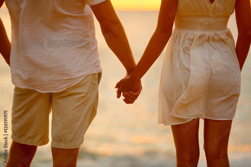 Romantic couple holding hands on beach sunset during travel. Closeup of happy woman and man together in romance on romantic honeymoon getaway in beautiful light. Casual beachwear white linen clothing. Romantic couple holding hands on beach sunset during travel. Closeup of happy woman and man together in romance on romantic honeymoon getaway in beautiful light. Casual beachwear white linen clothing.